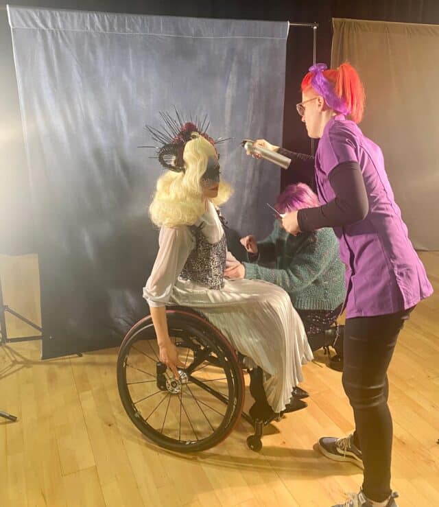 Blond haired woman in wheelchair in grey dress. Red haired woman in purple wearing purple top and applying hairspray