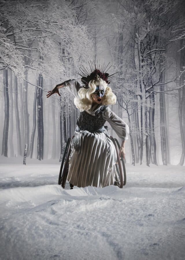 white snow covered trees with masked blonde haired woman in grey dress on a wheelchair