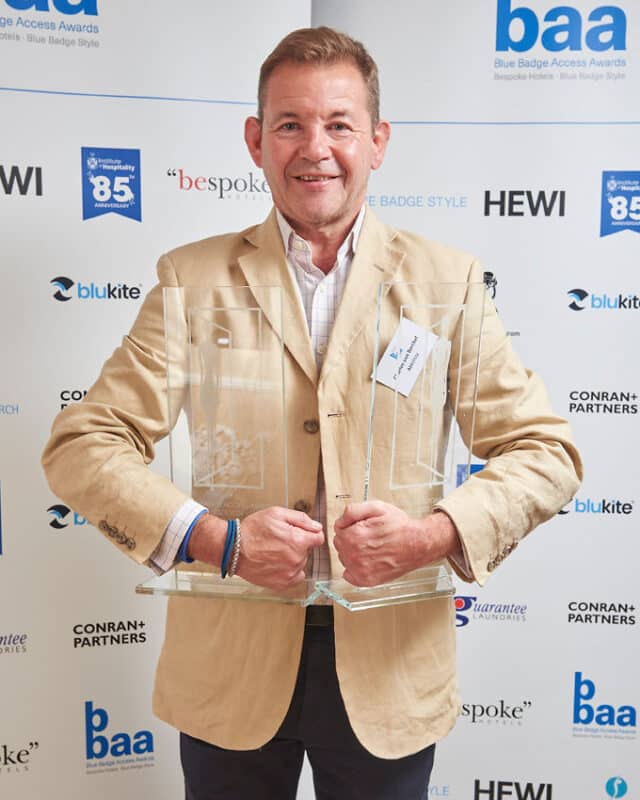 Man in cream jacket holding 2 glass awards