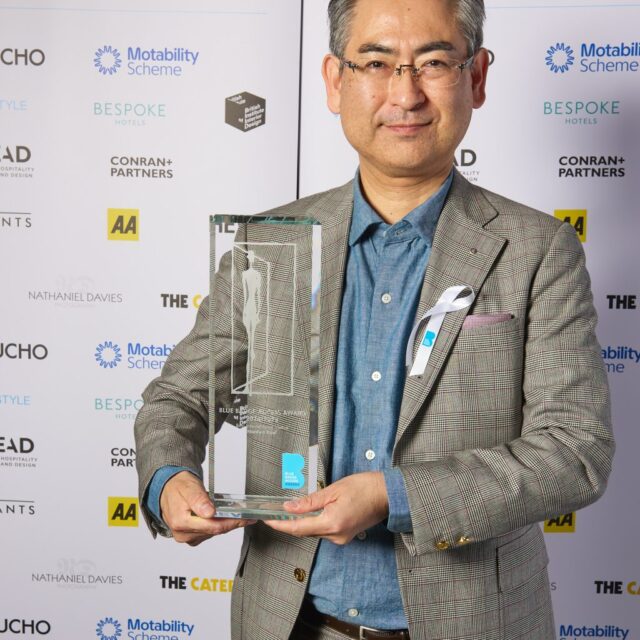 japanese man with grey hair wearing black & white suit with blue shirt holding glass award